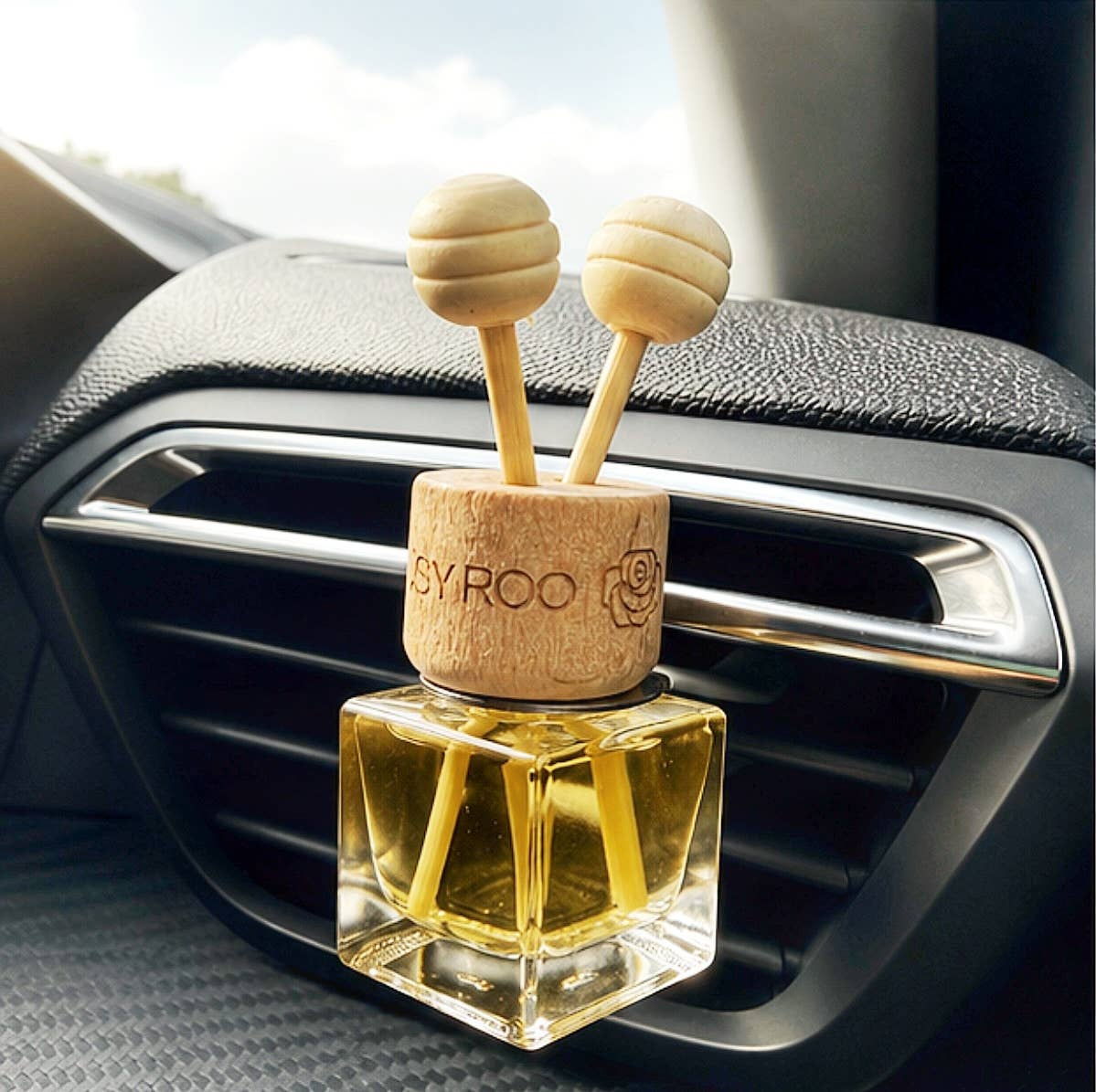 A car air freshener with a clear glass bottle filled with yellow oil and a natural wood cap featuring two round wooden diffuser tops, shown plugged into a car air vent.