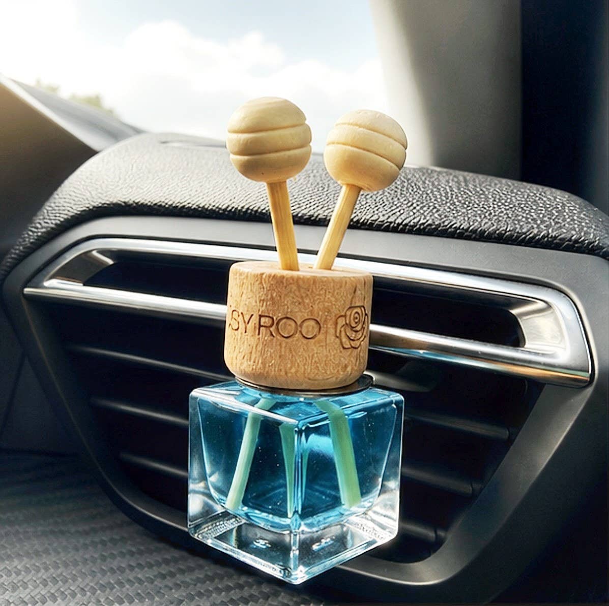A car air freshener diffuser with a clear blue glass bottle and two light wood spherical reeds, mounted on a car dashboard vent. The wooden top is engraved with the brand name 'BYROO'.