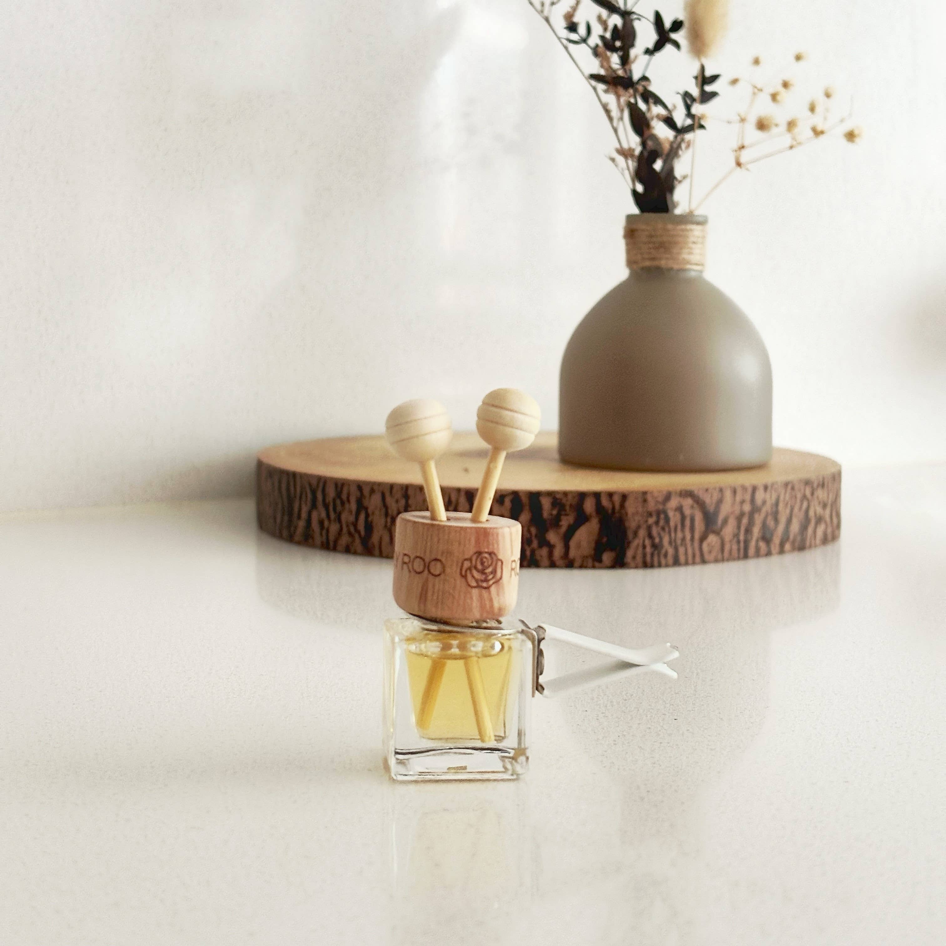 A small glass reed diffuser bottle with amber liquid and wooden diffuser sticks sits in front of a matte taupe vase holding dried stems, both displayed on a round wood slice against a soft white background.