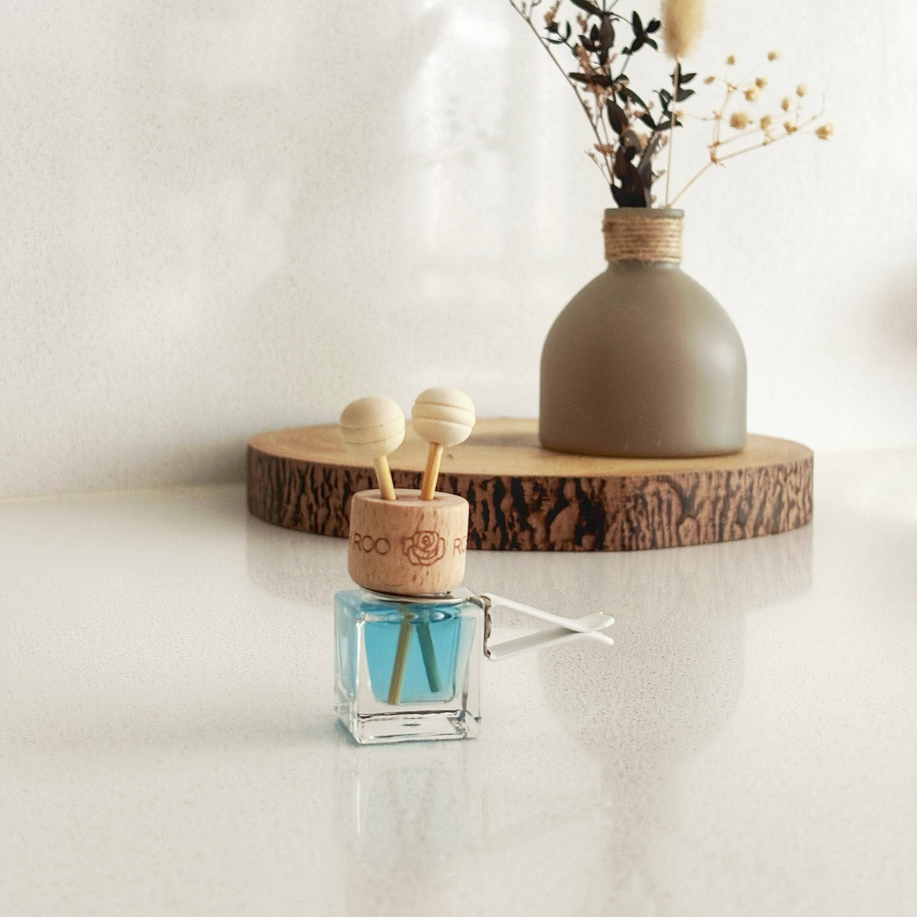 A small reed diffuser bottle with blue liquid and wooden diffuser sticks sits in the foreground on a white surface, while a matte taupe vase with dried stems rests on a round wood slice in the background against a soft white wall.