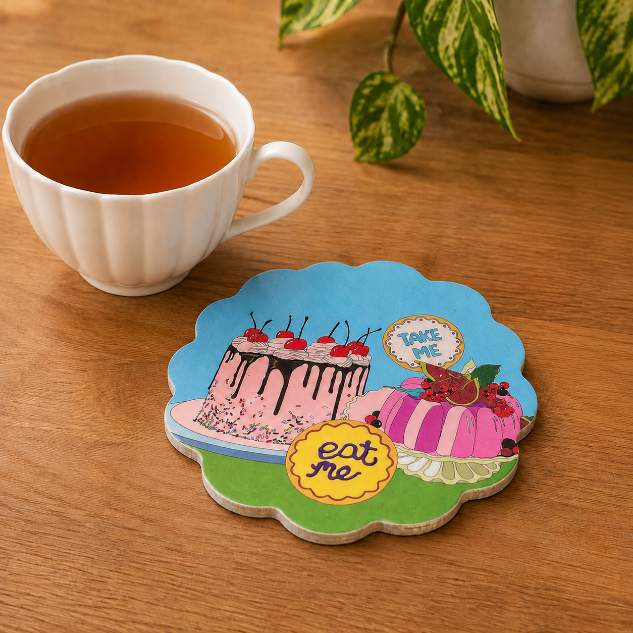 Alice in Wonderland tea party scallop coaster styled beside a cup of tea, featuring colourful cakes and Eat Me and Take Me details.