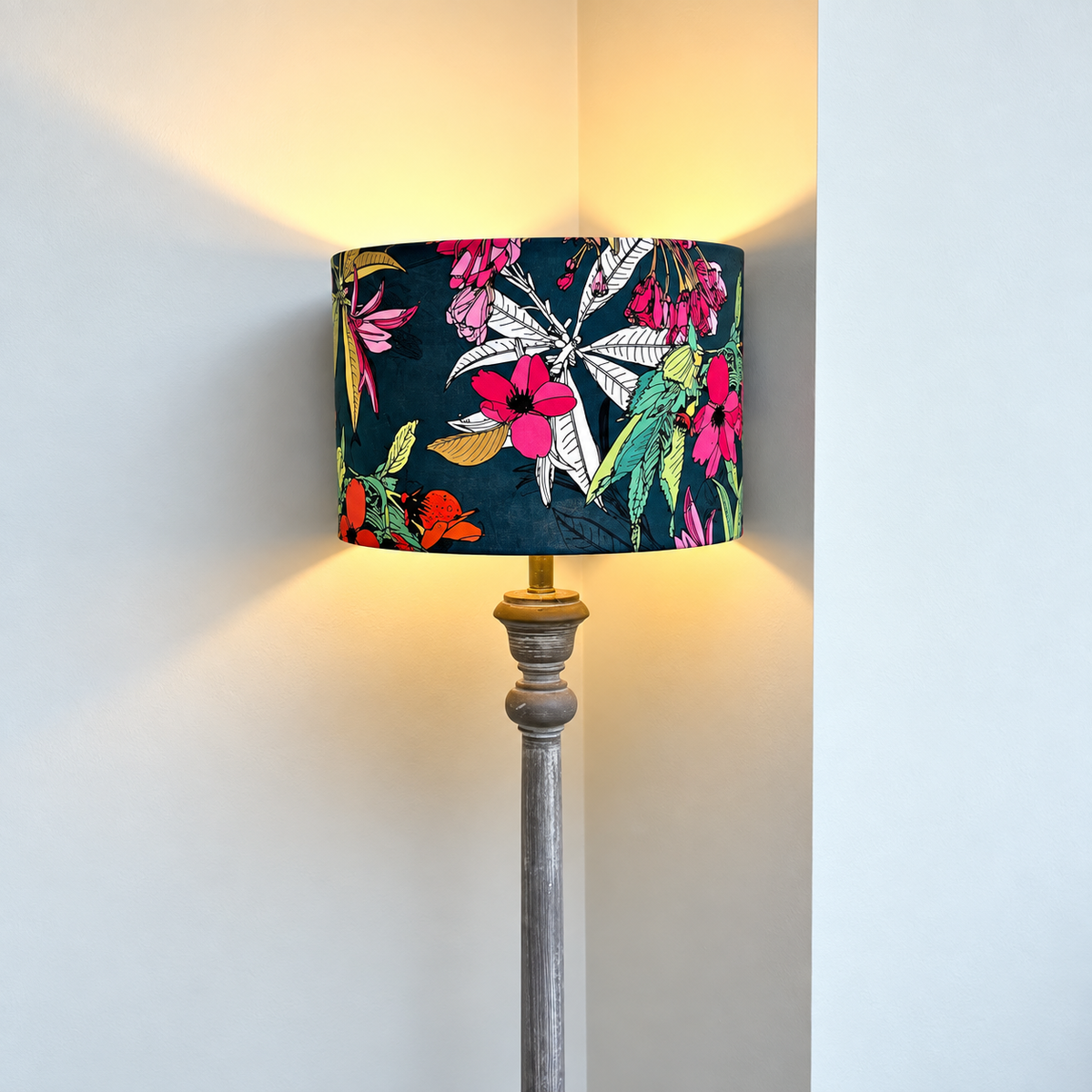 A floor lamp with a cylindrical drum shade featuring a vibrant tropical floral and bird pattern on a dark teal background, set against a plain light gray wall with warm light glowing from the lamp.