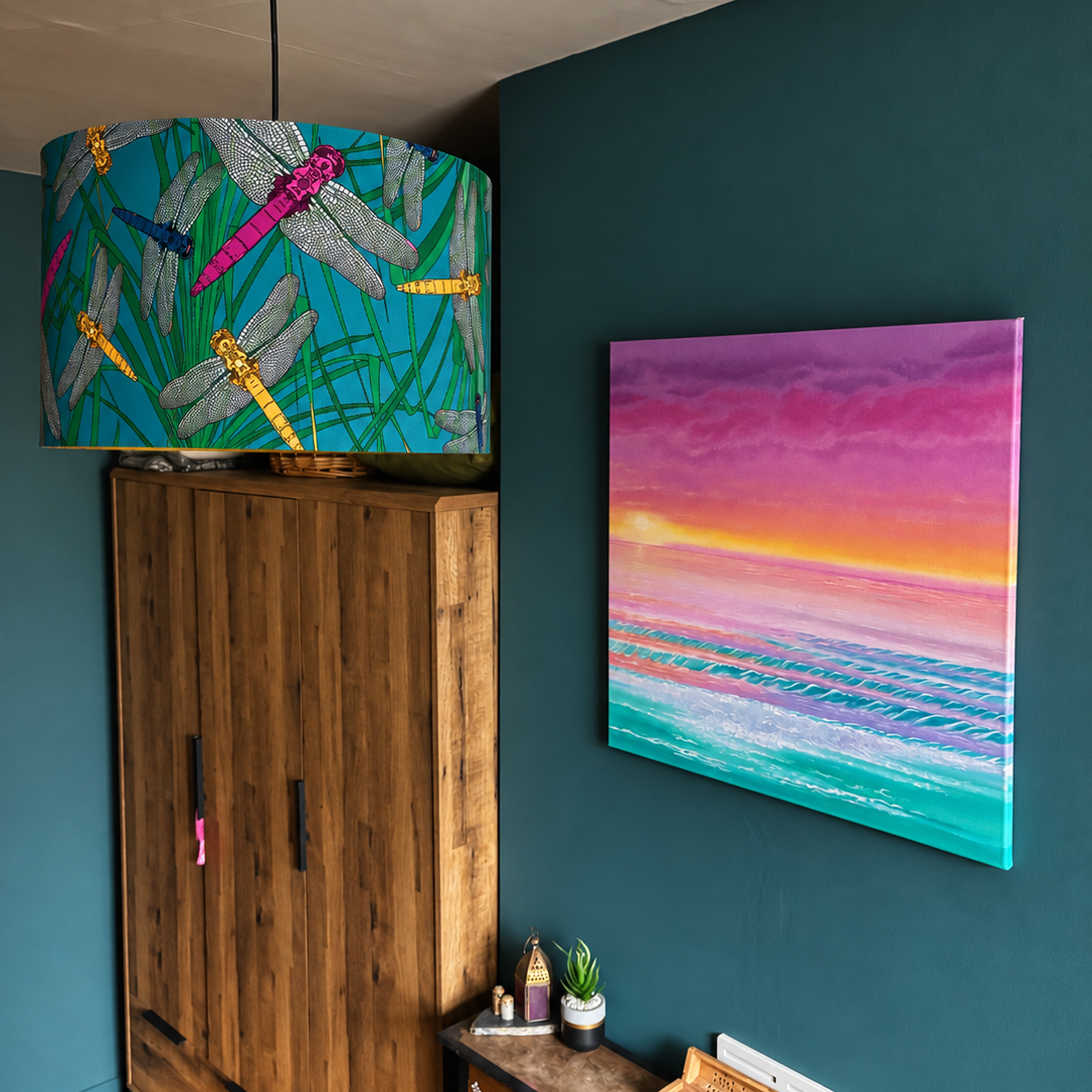 A colorful dragonfly-patterned drum pendant lamp hanging in a room with a dark teal wall, a wooden cabinet, and a framed sunset painting.