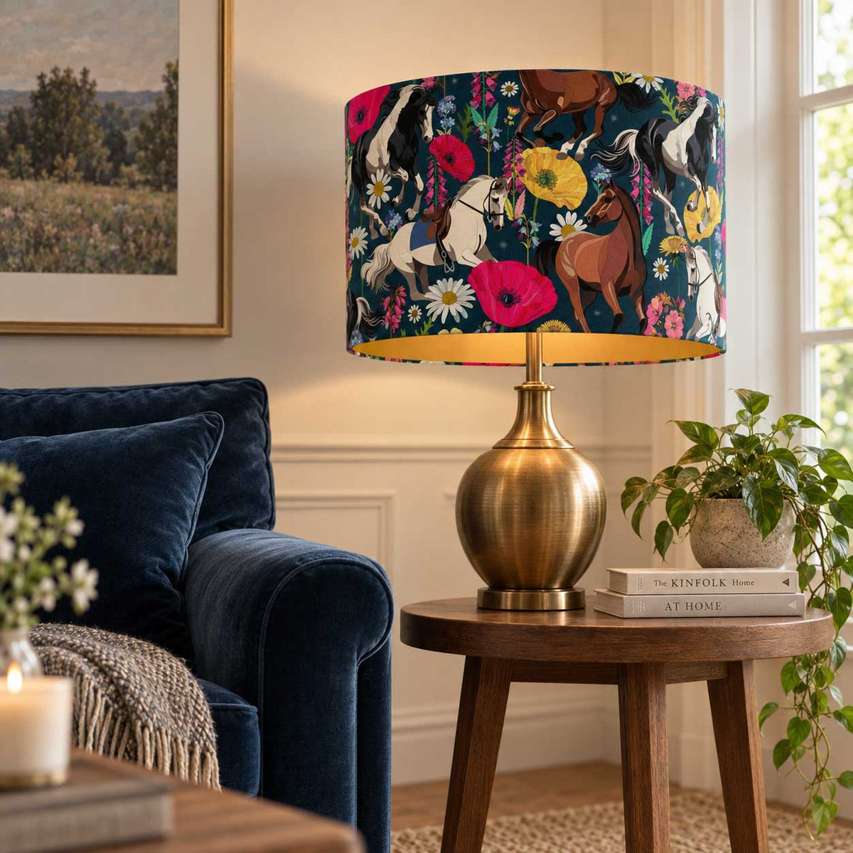A table lamp with a dark navy blue drum shade covered in a colorful horse and floral print, set on a round wooden side table in a cozy living room next to a blue velvet sofa.