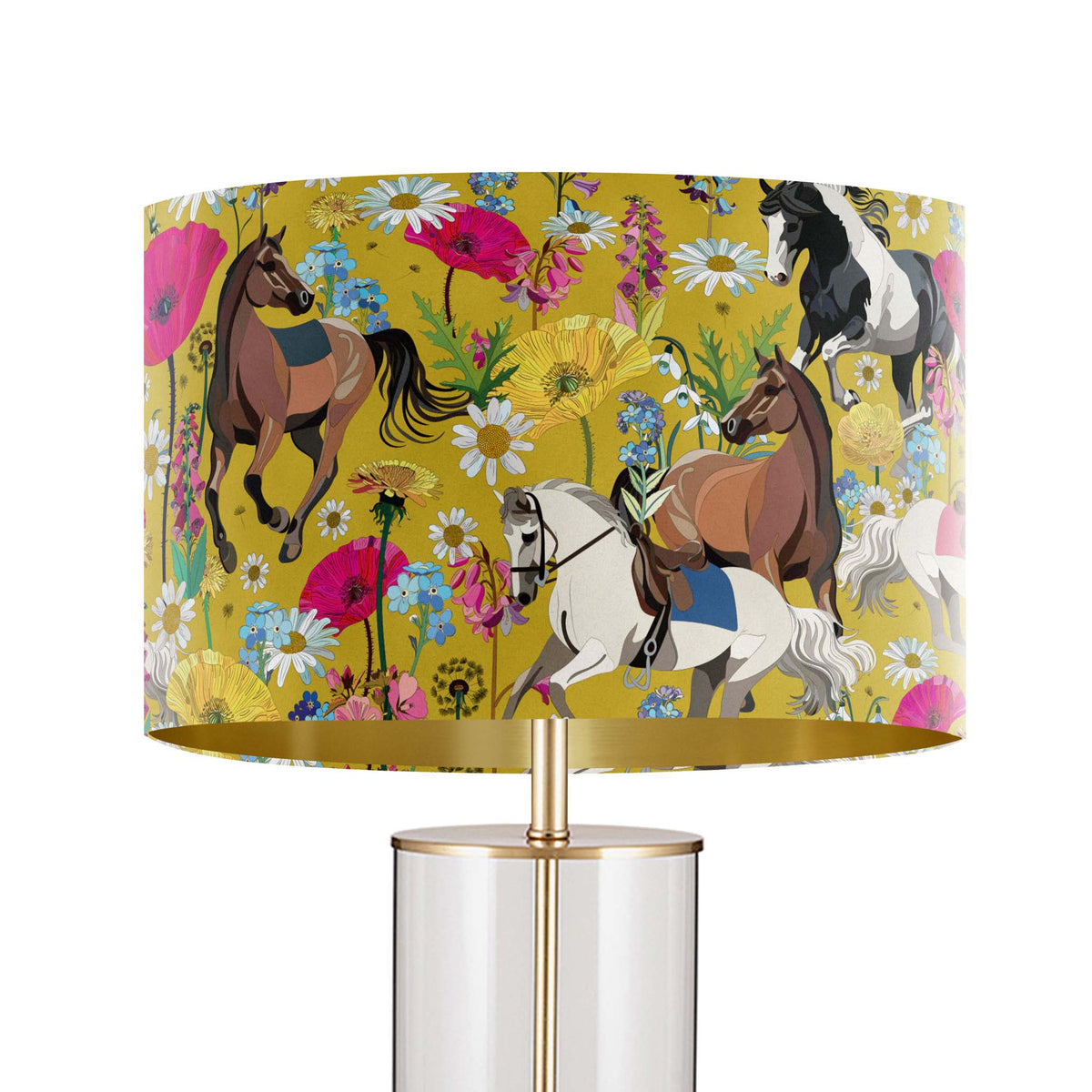 A table lamp with a cylindrical clear glass base and a gold-finished stem, topped with a wide drum shade featuring a colorful print of horses and wildflowers on a mustard yellow background.