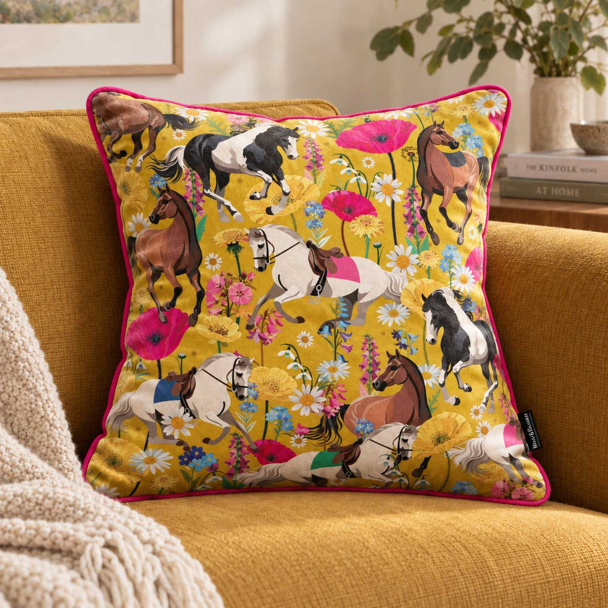 A square decorative throw pillow with a vibrant yellow background and a colorful print of horses, flowers, and poppies, styled on a mustard yellow armchair next to a cream knit blanket.