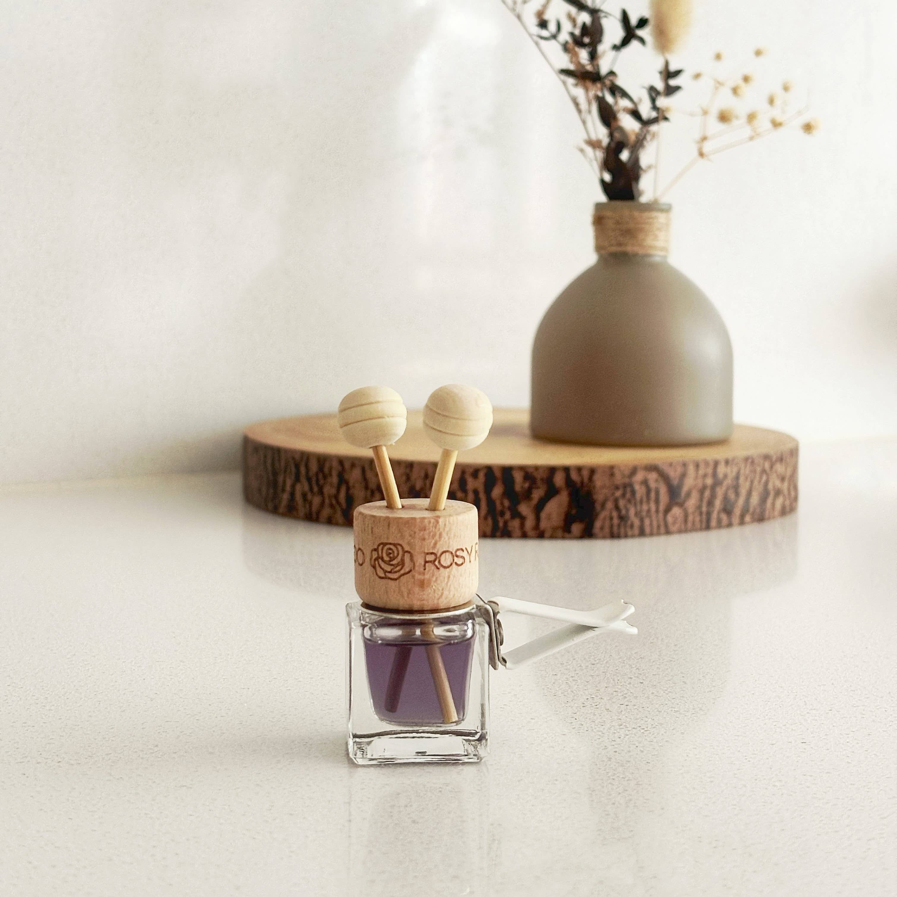 A car air freshener with a clear glass bottle filled with yellow oil and a natural wood cap featuring two round wooden diffuser tops, shown plugged into a car air vent.