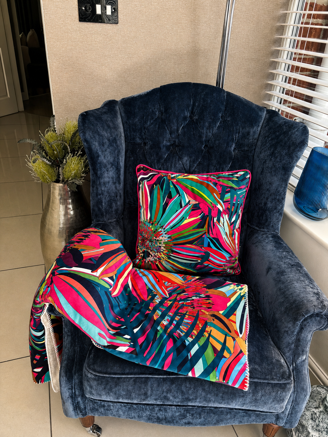 A navy blue velvet wingback armchair in a home setting, styled with a vibrant multicolor tropical palm leaf throw blanket and a matching square throw pillow.