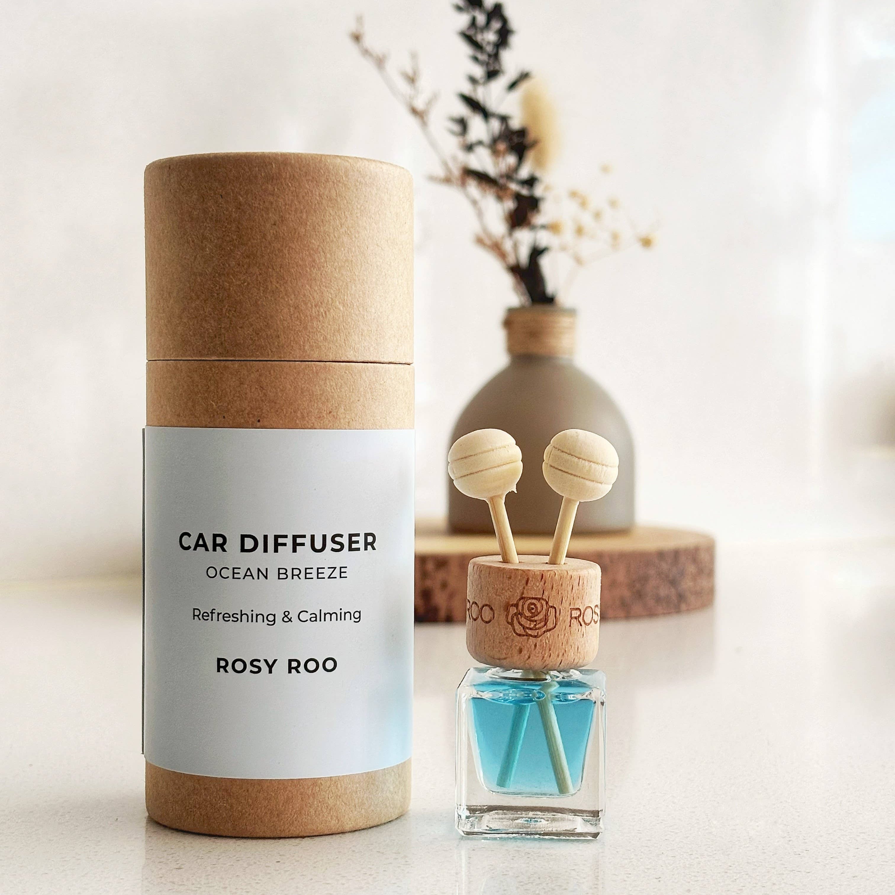 A car diffuser bottle with blue liquid and two wooden dropper tops stands beside its kraft paper cylindrical packaging on a light surface. The label reads 'CAR DIFFUSER OCEAN BREEZE Refreshing & Calming ROSY ROO'.