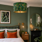 A styled bedroom interior featuring a green patterned drum pendant lamp with a gold interior, hanging above a bed with orange and green pillows in a dark green room.