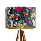 A tripod floor lamp with a dark teal drum shade featuring a colorful pattern of horses and wildflowers, finished with a gold inner rim and a wooden tripod base.