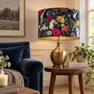 A table lamp with a dark navy blue drum shade covered in a colorful horse and floral print, set on a round wooden side table in a cozy living room next to a blue velvet sofa.