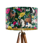 A tripod floor lamp with a dark green drum shade featuring a colorful pattern of horses and wildflowers, with a gold inner rim and a wooden tripod base.