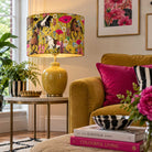 A yellow ceramic table lamp with a vibrant, multicolored horse and floral patterned drum shade, styled in a cozy living room next to a mustard yellow sofa and pink pillows.