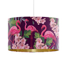 A drum pendant lampshade featuring a vibrant tropical print of pink flamingos, green leaves, and light pink blossoms on a deep purple background, finished with a shiny gold metallic inner rim.