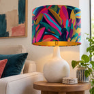 A table lamp with a colorful abstract leaf-patterned drum shade and a textured off-white ceramic base, styled on a wooden side table in a cozy living room setting.