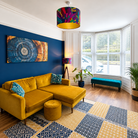 Colourful abstract velvet ceiling lampshade with rich pink, teal, green and purple tones and a gold inner, styled in a bright living room with a mustard velvet sofa, navy feature wall and matching table lamp.
