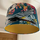 A fabric drum lampshade with a tropical bird and floral print in teal, gray, pink, yellow, and white, featuring a solid mustard yellow interior, shown hanging under a ceiling.