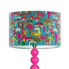 A table lamp with a bright pink beaded stem and a cylindrical drum shade covered in a colorful mushroom print on a turquoise background, isolated on a white background.