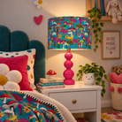 A colorful children's bedroom scene featuring a bright pink and turquoise mushroom-patterned table lamp on a white nightstand next to a teal velvet bed headboard and matching patterned bedding.