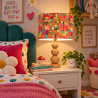 Orange mushroom velvet bedside lampshade with colourful hand-illustrated toadstools and flowers, styled on a natural wood lamp base in a playful children’s bedroom.