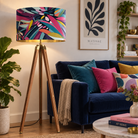 A tall floor lamp with a colourful abstract leaf-patterned drum shade and a wooden tripod base, styled in a cozy living room next to a blue velvet sofa with colourful pillows.