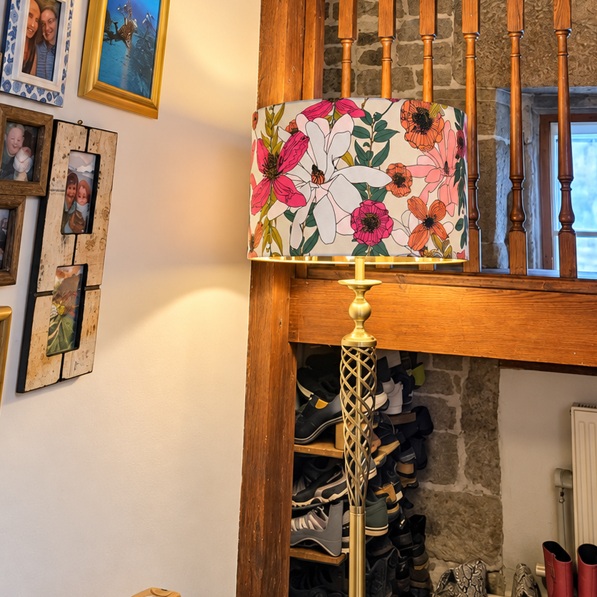 A floor lamp with a white drum shade covered in a vibrant pink, orange, and green floral print, supported by a gold-toned metal stand in a home interior next to a wooden staircase.