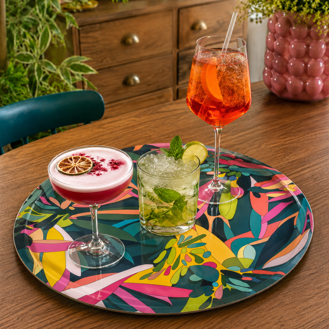 A round cocktail tray with a colourful teal abstract print, styled on a wooden table with three cocktails in glasses.