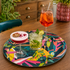 A round cocktail tray with a colourful teal abstract print, styled on a wooden table with three cocktails in glasses.