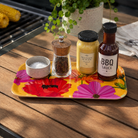 A rectangular yellow tray with a bold pink and red floral print sits on a wooden outdoor table. On the tray are a small salt bowl, a pepper grinder, a jar of mustard, and a bottle of BBQ sauce, with a potted plant and grill visible in the background.