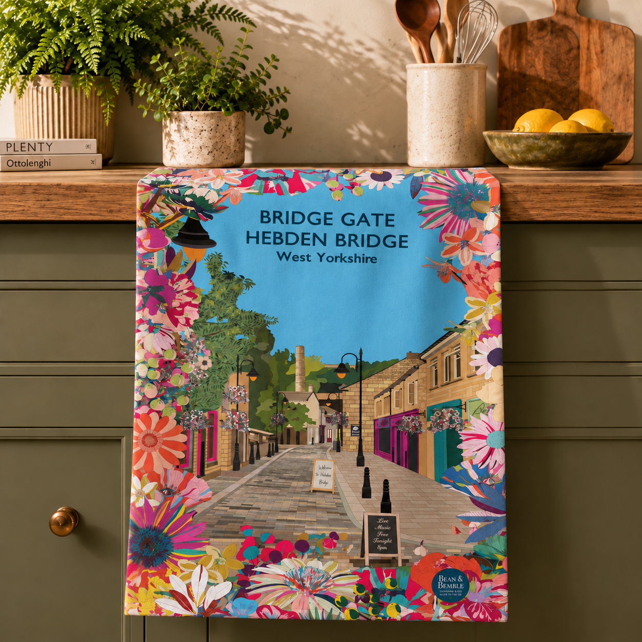 Colourful Hebden Bridge tea towel featuring Bridge Gate, stone buildings, independent shopfronts, hanging baskets and bright flowers.