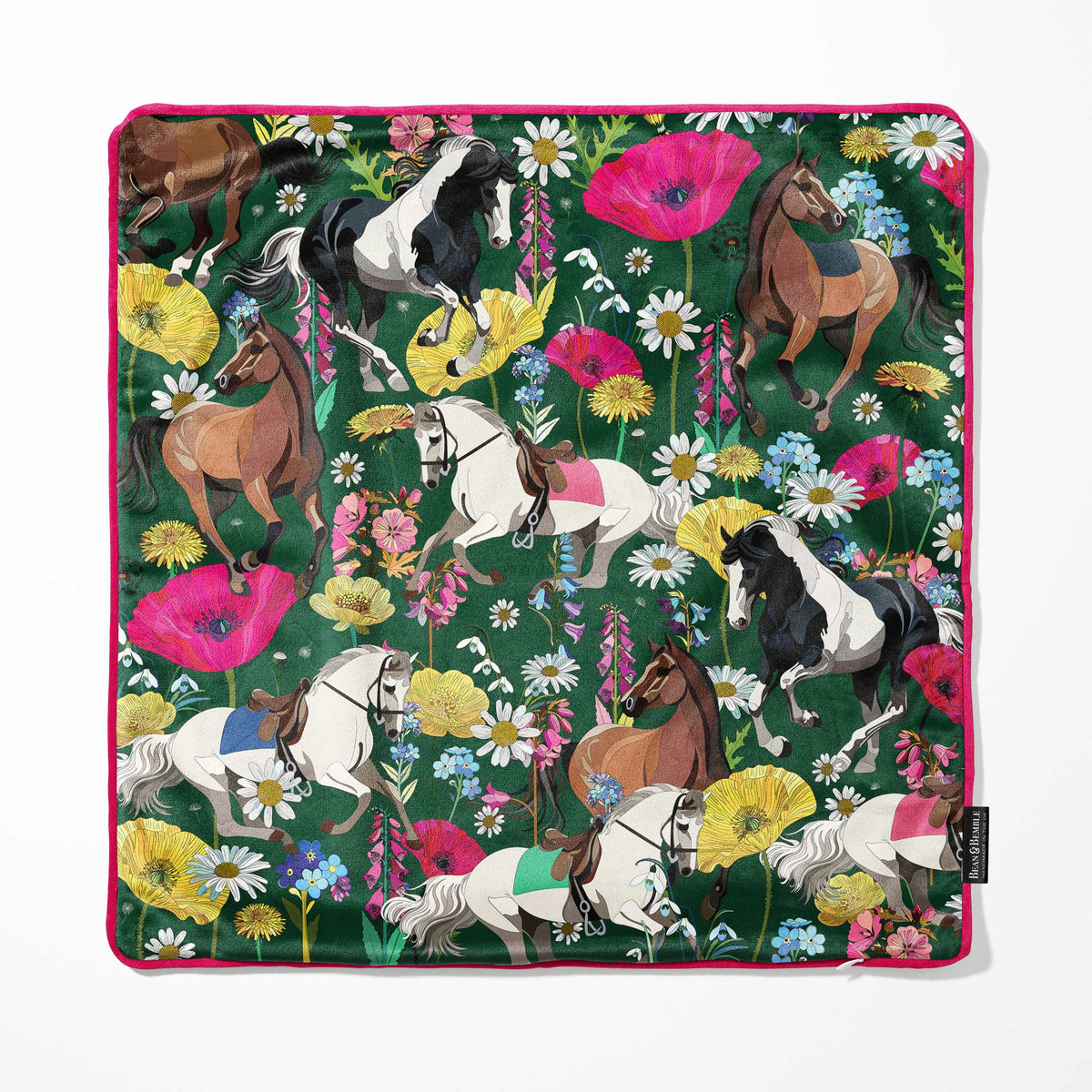 A square cushion cover with a vibrant floral and equestrian pattern, featuring illustrated horses and wildflowers on a dark green background with a bright pink piped edge. A small black Bean and Bemble brand tag is visible on the bottom right edge.