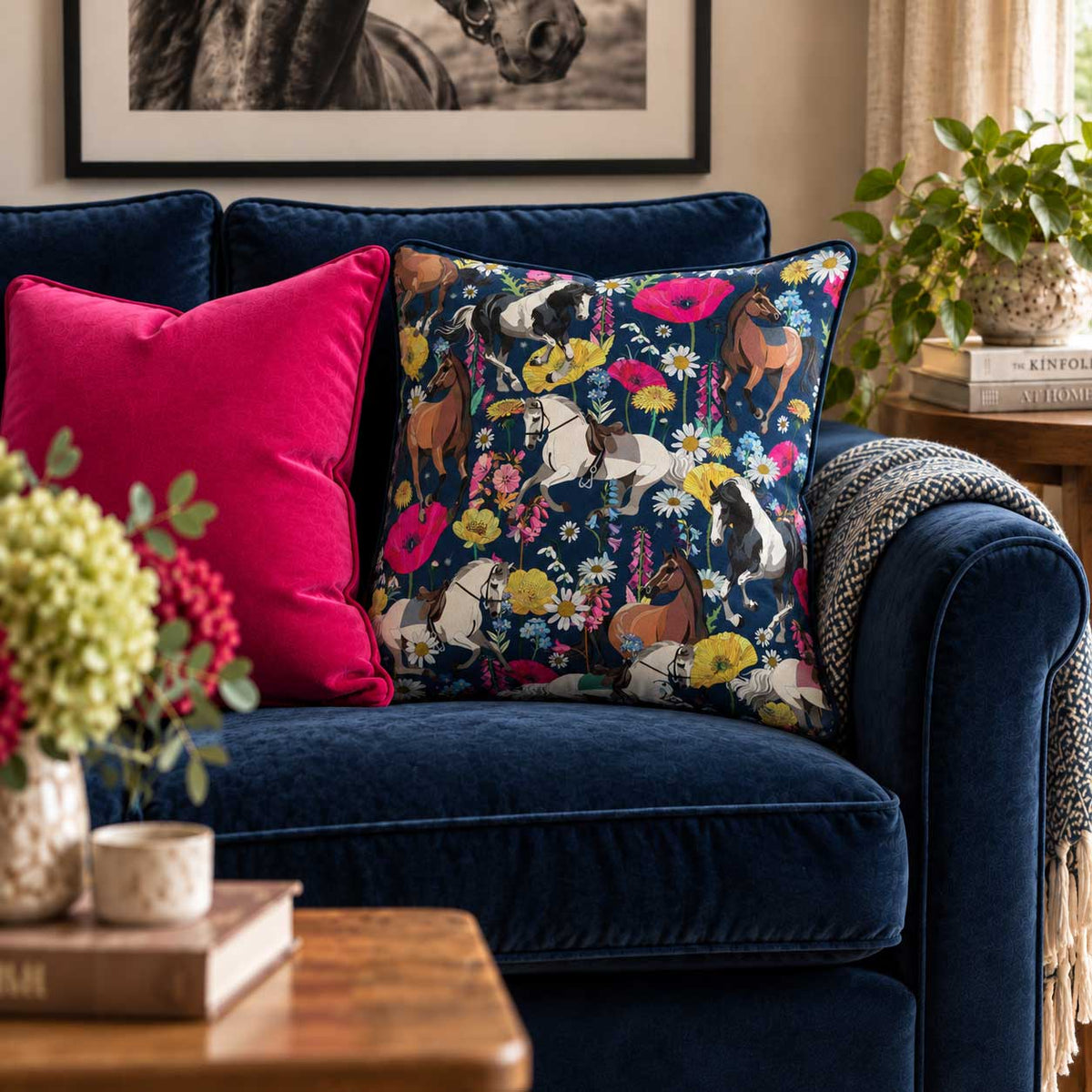 A navy blue throw pillow with a colourful horse and floral print is styled on a deep navy velvet sofa next to a solid hot pink pillow, in a cozy living room setting.