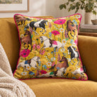 A square decorative throw pillow with a vibrant yellow background and a colorful print of horses, flowers, and poppies, styled on a mustard yellow armchair next to a cream knit blanket.